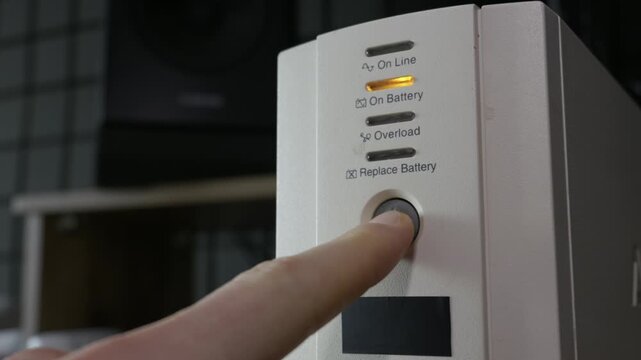 Close-up of a finger pressing the power button on a white UPS unit, activating the amber light. Concept of electrical safety, data protection, and office equipment.