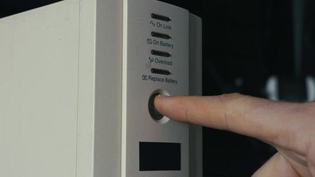 Finger presses power button on a white uninterruptible power supply unit, activating orange indicator light. Concept of emergency electricity, server maintenance, and hardware protection.
