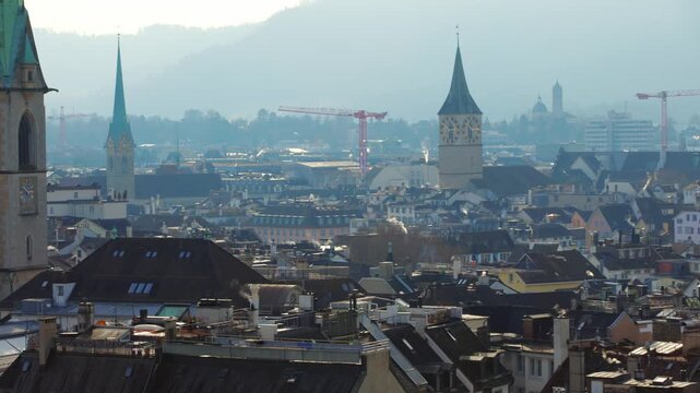 Wide panning shot over the historic rooftops of Zurich's old town, featuring the iconic church towers of Predigerkirche and St. Peter against a misty morning horizon in Switzerland.