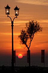 Obraz premium Vibrant orange sunset over the ocean featuring silhouetted Victorian street lamps, a lone tree, and red flags on shore.
