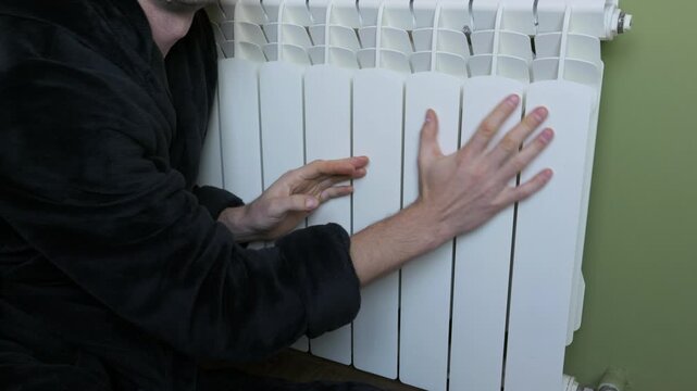 Man in a black bathrobe touches a white radiator to check the temperature inside a room. Concept of heating season, rising energy costs, and freezing winter weather problems.