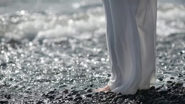 Woman standing barefoot on a seashore with sea waves caressing her bare feet. Closeup of her legs.