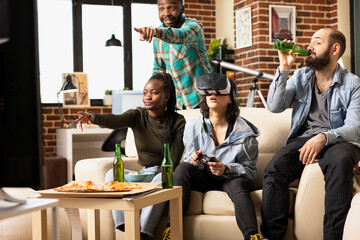 Caucasian woman uses virtual reality glasses and gamepad while playing online tournament, as diverse friends on couch watch. Female gamer with VR headset stays focused during casual apartment hangout. © DC Studio