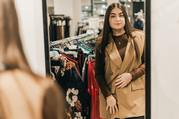 Young woman with long dark hair wearing a beige vest poses in front of a mirror inside a clothing...
