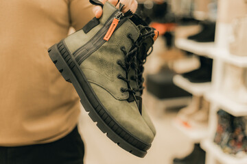 Person holding green hiking boots with black soles and orange zipper detail inside a shoe store, shelves of footwear visible in the background © Emvats
