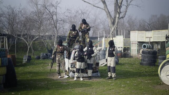 A group of paintball players in tactical gear and masks pose with their guns on an outdoor battleground. The team stands ready for action amidst car wrecks and obstacles in a strategic combat game.