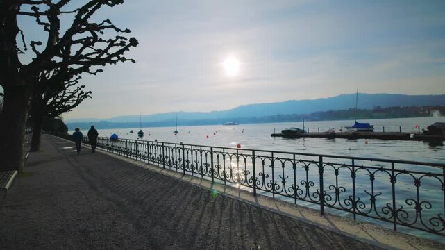 Static shot of the scenic Lake Zurich promenade at sunset, featuring a classic iron railing, silhouettes of people walking, and a calm lake view with boats and distant mountains.