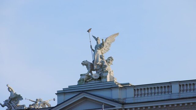 Panning shot of the ornate statues and classical sculptures atop the facade of the Zurich Opera House against a clear blue sky in Switzerland.
