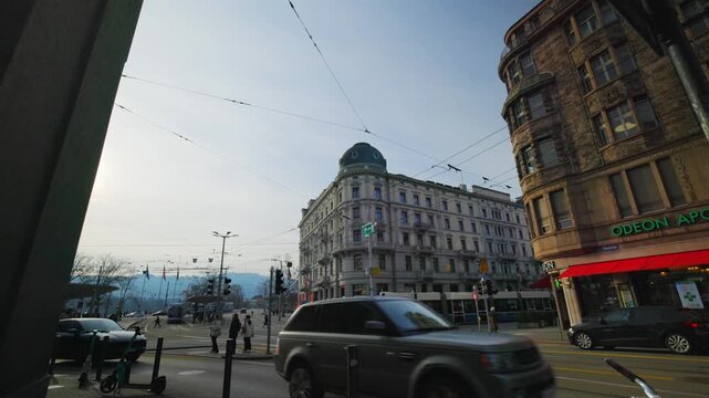 Static shot of the bustling Bellevue square in Zurich, Switzerland, featuring the historic Odeon building, street traffic, and overhead tram wires under a bright sky.