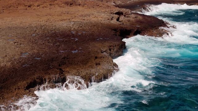 Impresionante vista a&eacute;rea de las olas rompiendo sobre las rocas rojizas de Aruba, revelando el hermoso paisaje con acantilados y las texturas de las rocas al sol