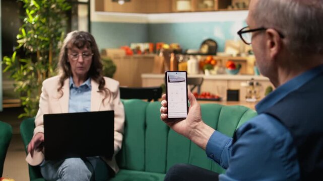 Aged man and woman reviewing green energy system through software on mobile phone. Sustainable, eco and clean power solutions like photovoltaic panels and off-grid solar energy at home.