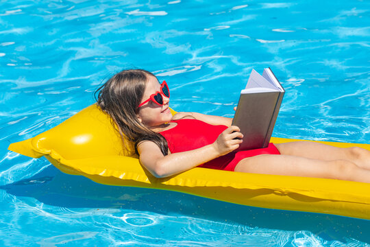 Side view little girl in red swimsuit relaxes on a yellow float in a swimming pool while reading a book under bright sunlight. Tropical vacation concept. High quality photo. Copy space