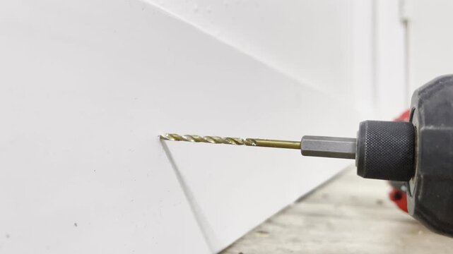 Cordless power drill with long masonry bit drilling into white drywall near baseboard home renovation and diy installation concept for construction repair carpentry and handyman work
