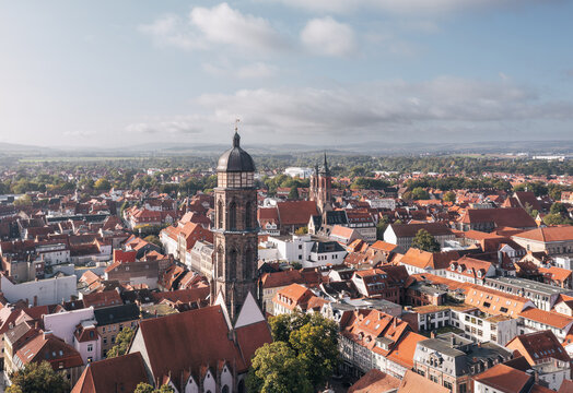Drone cityscape of Goettingen (G&ouml;ttingen), Germany, with a historic church tower rising above red tiled roofs and the old town stretching toward the horizon under a bright sky with clouds.