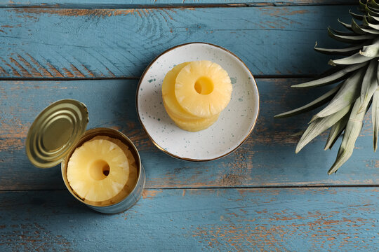 Plate and tin can with canned pineapple rings on blue wooden background