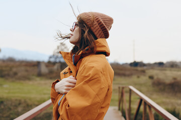 Young woman in orange jacket and brown knit hat stands outdoors on a bridge, enjoying fresh air and...