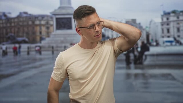 Man with glasses touching his head with right hand in a wet city street by a monument and buildings; thoughtful introspection.