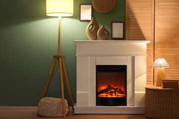 Naklejka premium Interior of evening living room with folding screen, glowing lamps, wicker table and home decor on fireplace near blue wall