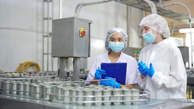 Female workers inspecting canned food product on automated conveyor belt production line inside food processing factory, quality control and hygiene inspection in manufacturing and safety compliance.