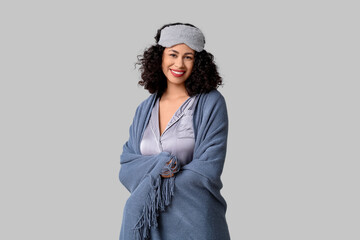 Beautiful young African-American woman in pajamas with cozy blanket and sleep mask on grey background © Pixel-Shot