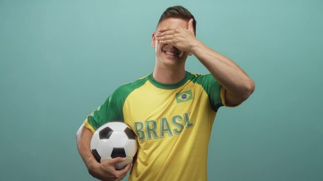 Young blond man in yellow brasil jersey with flag patch holding soccer ball, smiling, covers eyes with hand in studio; playful pride.
