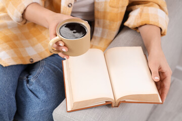 Obraz premium Young woman holding cup of hot coffee and book with blank pages on sofa at home, closeup