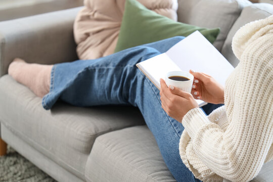 Young woman holding cup of hot coffee and book with blank pages sitting on sofa in living room