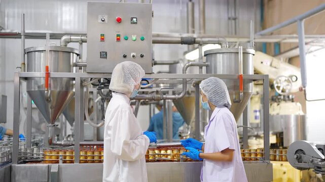 Female factory worker monitoring canned seafood production line and inspecting automated food processing machinery in manufacturing facility, quality control and industrial hygiene compliance workflow