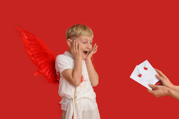 Naklejka premium Cute little shocked boy dressed as cupid with arrow and hands holding love letters on red background. Valentine's Day celebration