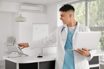 Fototapeta premium Male medical intern with laptop taking phone at reception in clinic