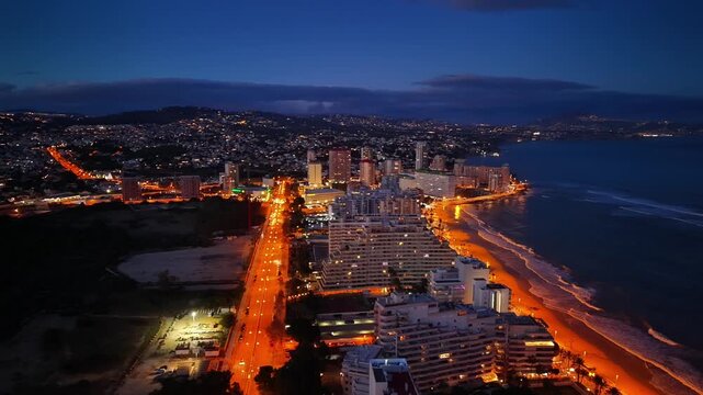 twilight aerial coastal city avenue lit by streetlights, predawn blue sky, orange road ribbon along beach,