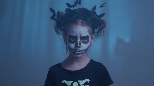 Little girl wearing skeleton face paint and dark costume surrounded by smoke. Portrait of child looking at camera with scary expression creating spooky Halloween atmosphere and dramatic mood