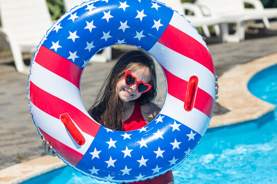 Happy smiling little girl in red swimsuit and sunglasses stands in outdoor swimming pool, holding a lifebuoy decorated with American flag print. Vacation in USA concept. 4th of July pool party