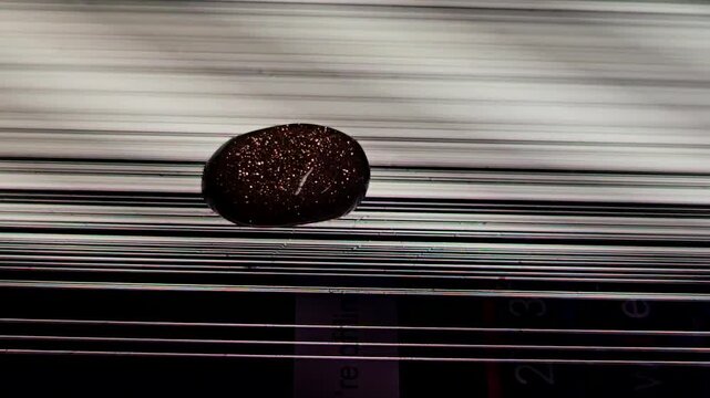 Close up of a dark smooth pebble on a polished metallic surface with sharp linear reflections.