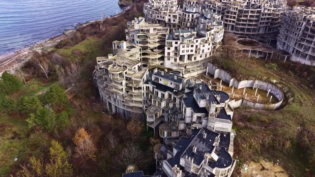 Crumbling concrete structures rise against the backdrop of a tranquil waterway, showcasing nature's reclamation of a once-bustling space. Trees surround the ruins, adding a touch of life.