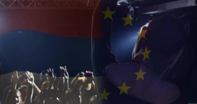Singer at gig raising, singing into mic to engage crowd filming phones, flag over singer silhouette