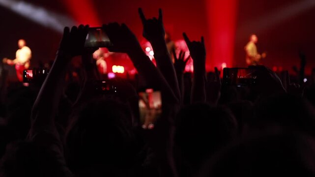 Fans cheer and make rock hand gestures during a live musical performance. The scene is illuminated by intense red and white stage lighting.