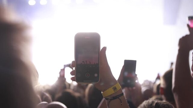 A fan films a musical event using a smartphone held above the crowd. The stage is brightly lit with white and red lights in a dark venue.