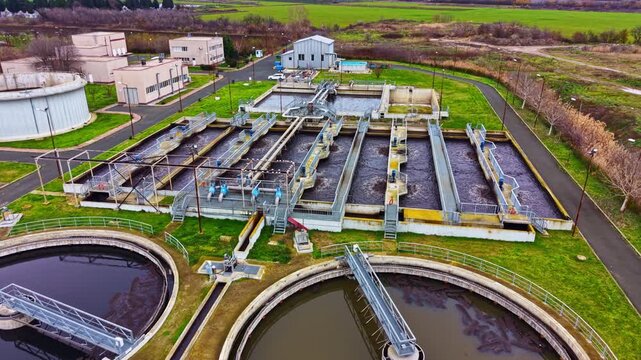 A water treatment facility shows large tanks and processing units. Pipes and machinery are visible. The site is surrounded by open land and trees. The day appears clear.
