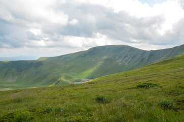 Rolling green hills and a gentle valley stretch beneath a cloudy sky. A winding path leads toward a small lake, inviting exploration and calm. Carpathian Mountains, Ukraine