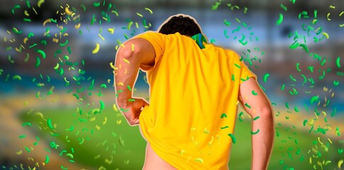 A man with his back to the camera, standing in the stands during his team's game, wearing yellow clothing with the grass in front of him, the stadium is full of fans, a young man cheering for his team © Cristiano Costa