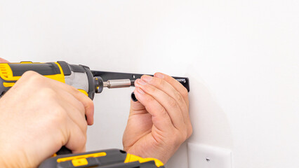 Mans hands tightening screw on black metal shelf bracket attached to white wall with cordless drill. Home improvement, final tightening, DIY shelf installation completion, secure fixture.