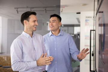 Fototapeta premium Collaborative project discussion. Two happy businessmen colleagues of diverse age ethnic backgrounds stand near wide digital display in corporate office share feedback enjoy successful sales results
