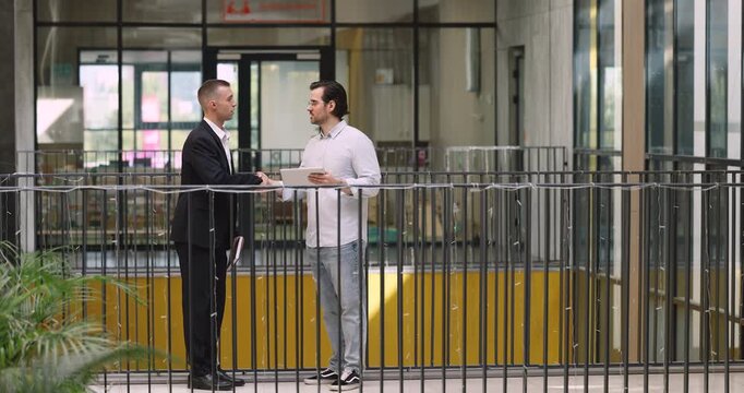 Two businessmen standing in corporate office hallway and reviewing online project or digital data on tablet, finalizing discussion with handshake gesture. Professional business collaboration, respect