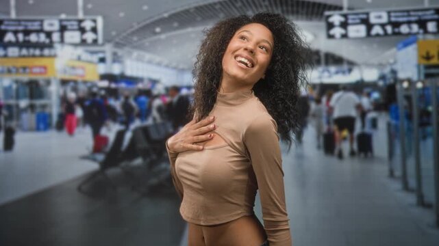 Teenage woman with curly hair places hand on chest in surprise, cropped top exposing bare midriff in an airport; joyful excitement and anticipation.