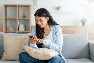 Obraz premium Smiling pretty young Asian woman sitting on cozy couch, using funny mobile apps in living room. Woman at Home, Doing Online Shopping, Messaging Friends, Posting on Social Media