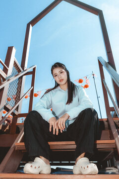 Confident young Asian woman in casual fashion clothing sitting on wooden staircase outdoor. low angle view of stylish person posing under clear blue sky, showing attitude