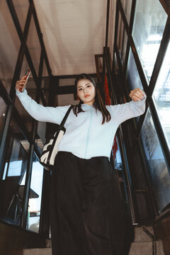 Confident young Asian woman in casual fashion takes selfie with smartphone while standing on staircase. This low angle shot shows her looking down with happy expression