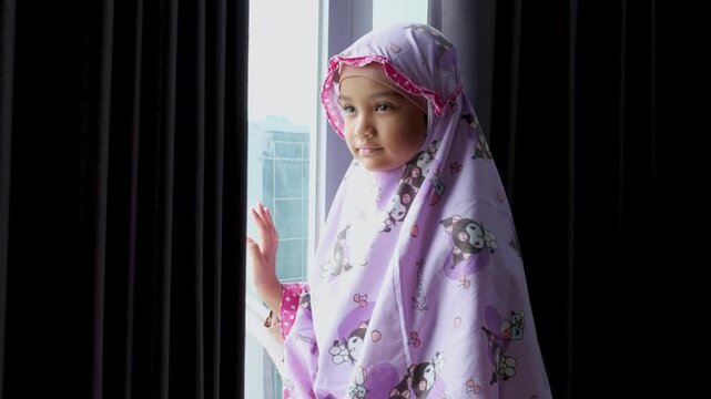 A beautiful Muslim girl wearing a prayer robe after praying is standing beside the window.