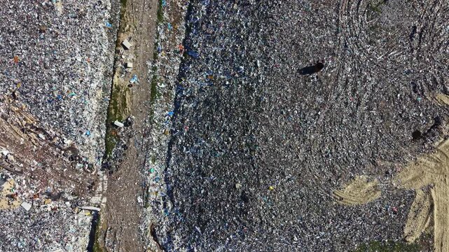 A large landfill site is spread across a rural field with various types of waste scattered all over. Machinery is present, working to manage the waste. The scene shows the impact of human activity.